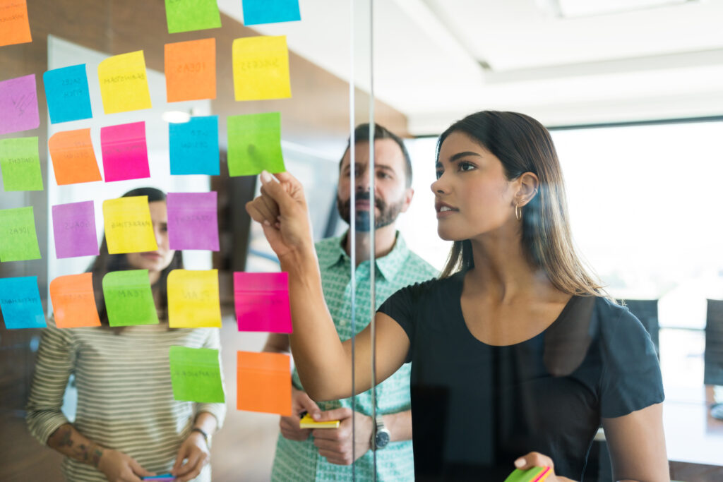 Drei Personen in einem modernen Büro in einem agilen Workshop-Setting. Im Vordergrund klebt eine Frau mit dunklen Haaren in schwarzem Oberteil bunte Post-its (gelb, pink, grün, blau, orange) an eine Glaswand und schreibt oder verschiebt Notizen. Hinter der Glaswand stehen zwei weitere Teammitglieder und beobachten bzw. diskutieren den Prozess. Die Szene wirkt kollaborativ und iterativ, typisch für agile Arbeitsmethoden wie Kanban oder Design Thinking. Die visuelle Trennung durch die Glaswand symbolisiert Transparenz und gleichzeitig Struktur im Arbeitsprozess. Die Post-its stehen für flexible Aufgaben, Ideen und sich entwickelnde Workflows.