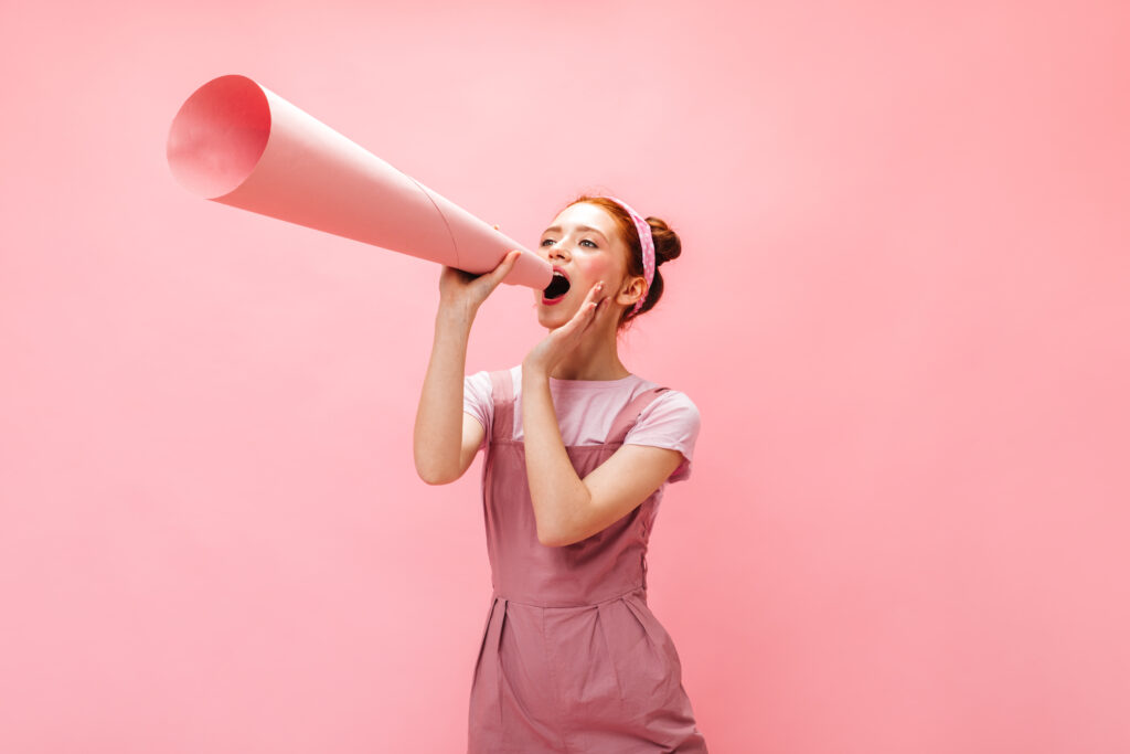 Eine Frau spricht durch ein großes Megafon vor rosa Hintergrund. Das Bild symbolisiert strategische Unternehmenskommunikation, Markenaufbau, Reputationsmanagement sowie die gezielte Vermittlung von Botschaften an Mitarbeitende, Kunden und andere Stakeholder.