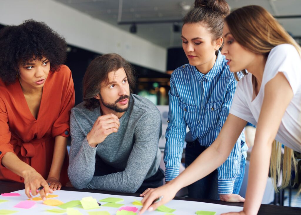 Interdisziplinäres Team arbeitet gemeinsam an einer agilen Lösung und organisiert Aufgaben mit Haftnotizen auf einem Tisch. Das Bild symbolisiert agile Transformation, Zusammenarbeit, Selbstorganisation, kontinuierliche Verbesserung und die Anpassungsfähigkeit von Unternehmen in einem dynamischen Umfeld.
